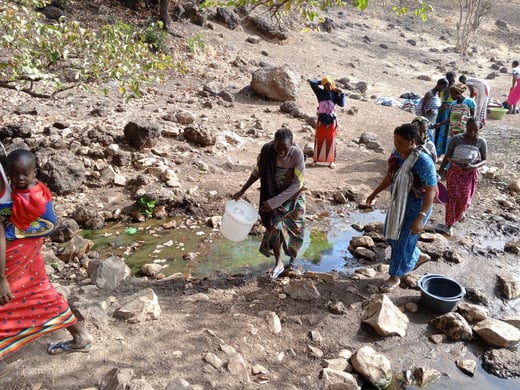 People collecting water with containers from a small stream in a rocky, arid area.