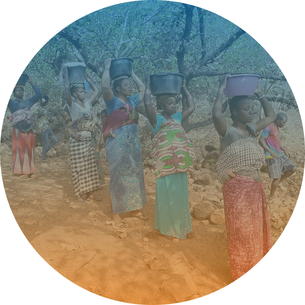 A group of women carrying containers on their heads walk along a dirt path outdoors, with trees and rocks in the background.