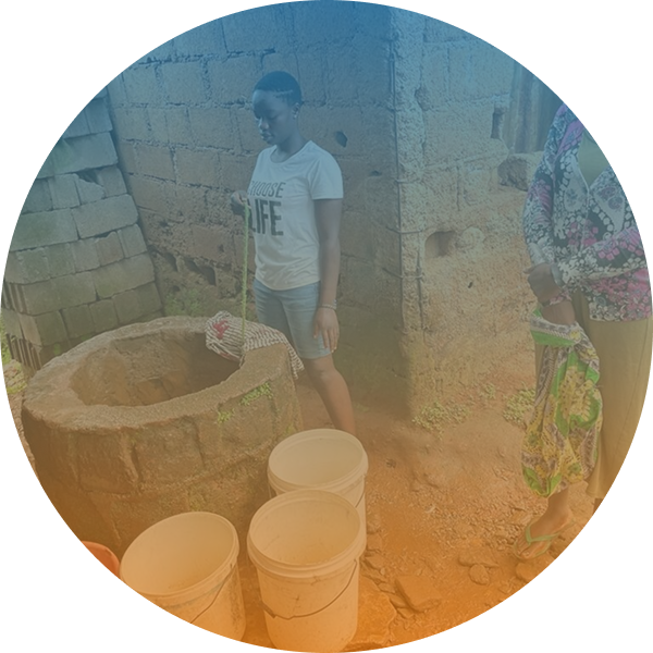 Two people stand beside a stone well with several empty buckets on the ground, next to a brick building in an outdoor setting.