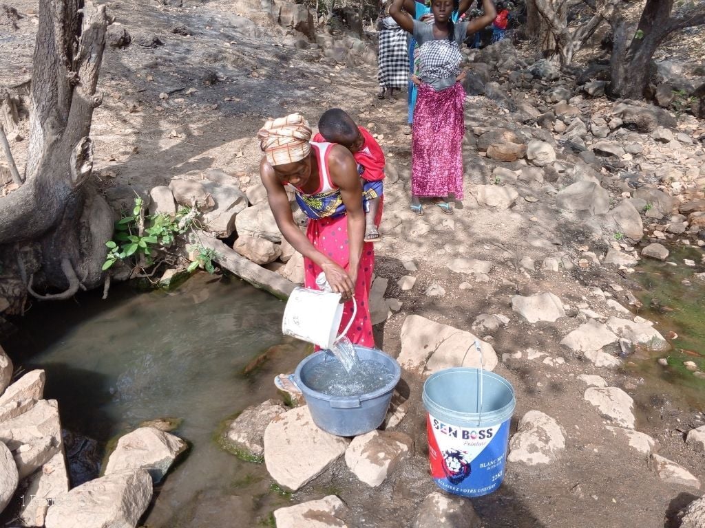 A woman with a child on her back fills a bucket with water from a stream using a plastic container; another woman stands in the background. Rocky, dry landscape surrounds them.