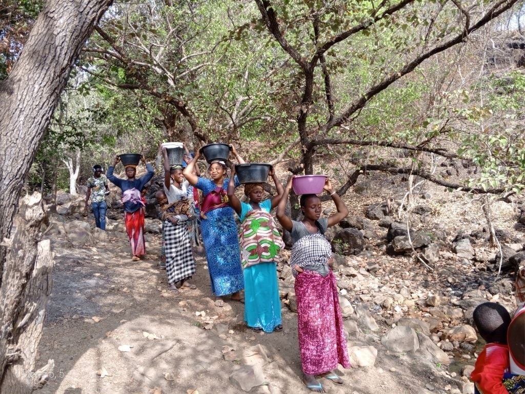 A group of women and girls walk along a rocky path carrying containers of water on their heads, surrounded by trees and dry terrain.