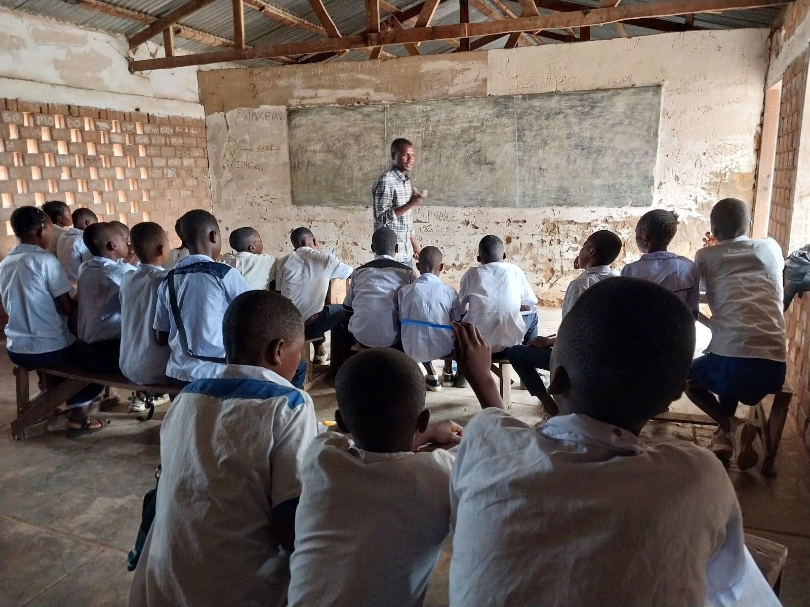 A teacher stands at the front of a classroom, instructing a group of students sitting on benches facing a chalkboard in a rustic, unfinished classroom.