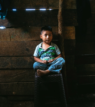A young boy sits on a small stool in a dimly lit wooden room. He is barefoot and wearing jeans and a green patterned shirt.